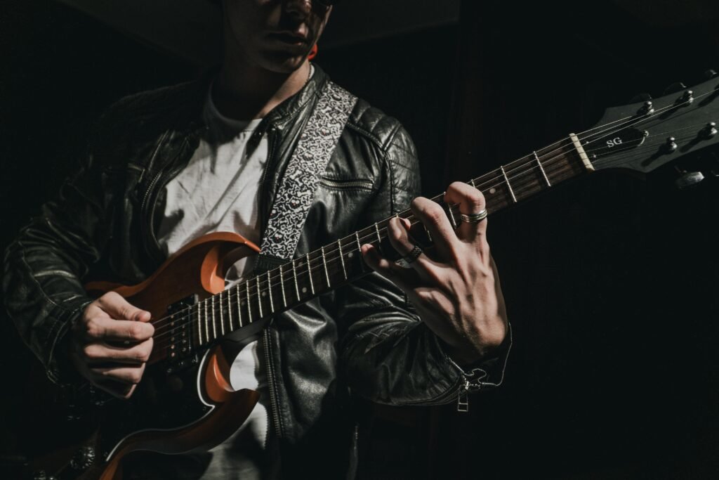 pexels-photo-33923310-33923310 Close-up of a guitarist strumming an electric guitar in a dark setting, wearing a leather jacket.